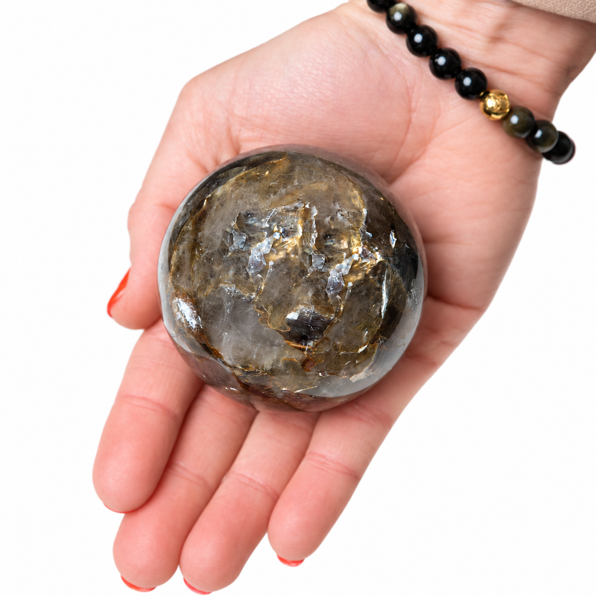 A hand holding a polished rough crystal sphere with natural gold and dark brown inclusions, shown against a plain white background. The hand also wears a black beaded bracelet with a small gold accent.