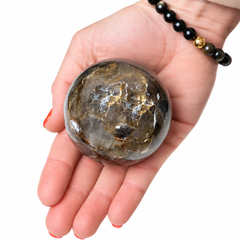 A hand holding a polished rough crystal sphere with natural gold and dark brown inclusions, shown against a plain white background. The hand also wears a black beaded bracelet with a small gold accent.