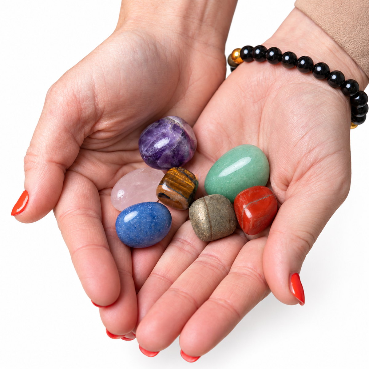 A pair of hands holding a collection of polished tumbled crystals, including purple amethyst, green aventurine, blue spiny oyster, pink rose quartz, brown tiger's eye, and red jasper, with a black beaded bracelet on one wrist, set against a solid white background.