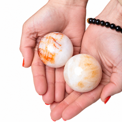 Two polished white stone spheres with orange and beige marbling are held in a person's hands against a white background. The person also wears a black beaded bracelet.