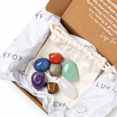 A set of seven polished gemstone crystals in various colors, arranged on a cream-colored drawstring pouch inside an open brown cardboard gift box lined with branded white tissue paper. The crystals include blue, red, green, purple, brown, and white stones.