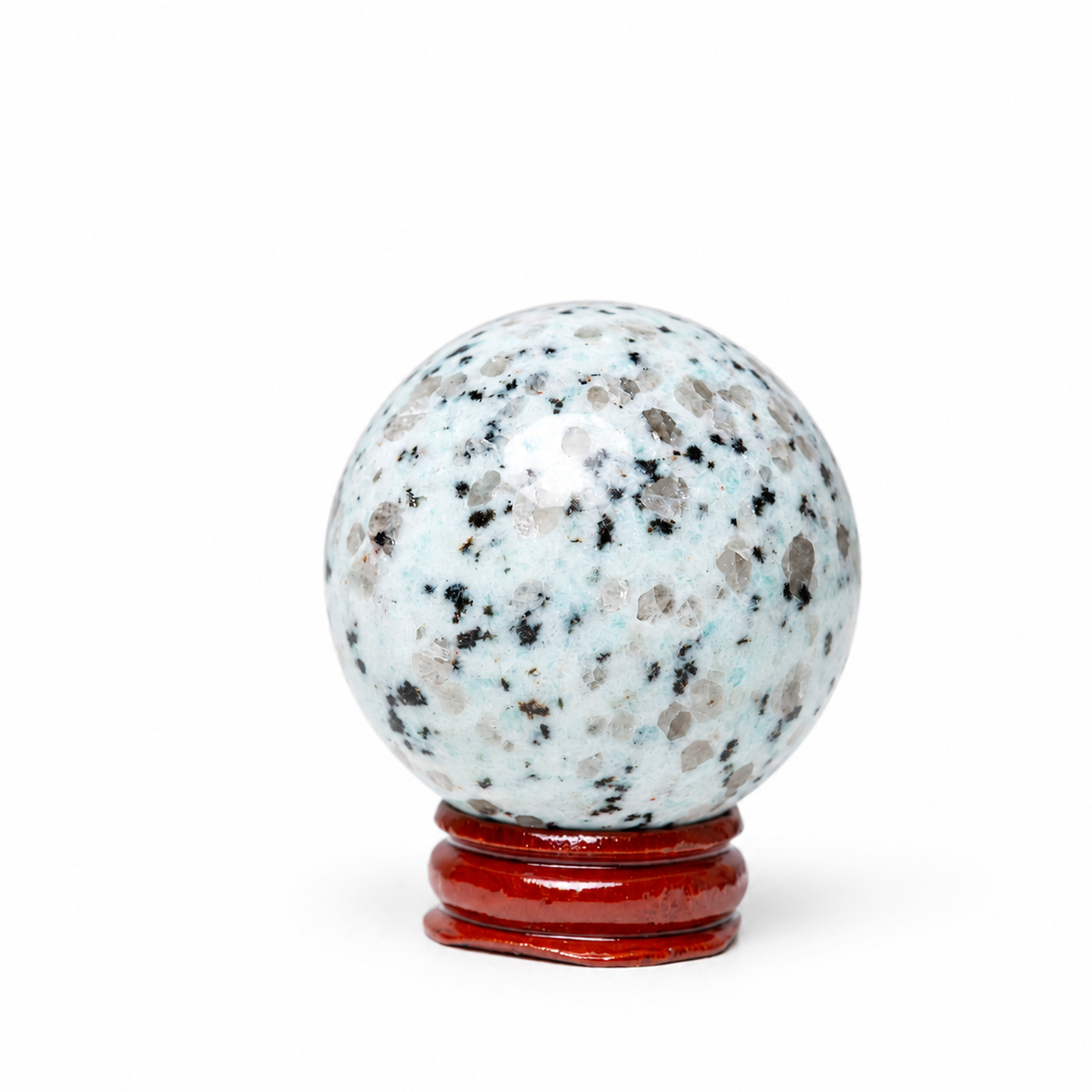 A polished crystal sphere with a light blue and white base speckled with black, gray, and brown flecks, displayed on a small red wooden stand against a plain white background.