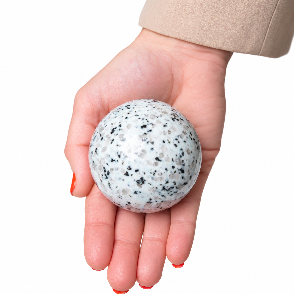 A hand with red nail polish holding a polished white stone sphere speckled with black and gray flecks, set against a plain white background.