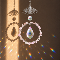 A decorative hanging ornament with a silver butterfly-shaped top, a circular ring of Kunzite stones, and a large iridescent teardrop crystal in the center, shown against a warm brown background with sunlight and rainbow reflections.