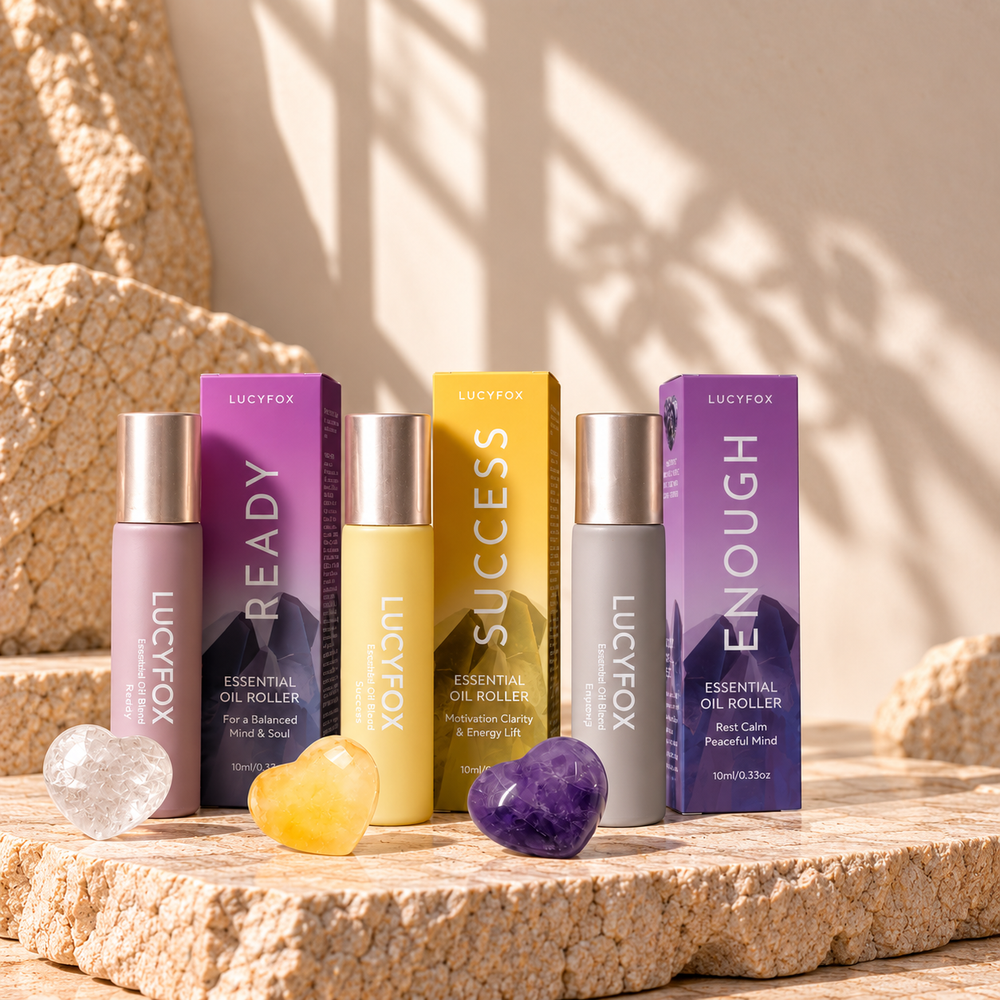 Four LUCYFOX essential oil roller bottles and matching boxes arranged on a stone slab with crystal balls. The products include Ready (lavender), Success (yellow), and Enough (purple) essential oil rollers, with their corresponding branded boxes behind them.