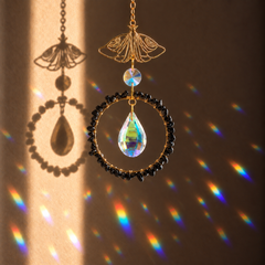 A decorative hanging ornament with a gold-tone butterfly-shaped top, a circular frame of small black beads, and a teardrop-shaped iridescent obsidian crystal pendant, shown against a warm brown wall with rainbow light projections.