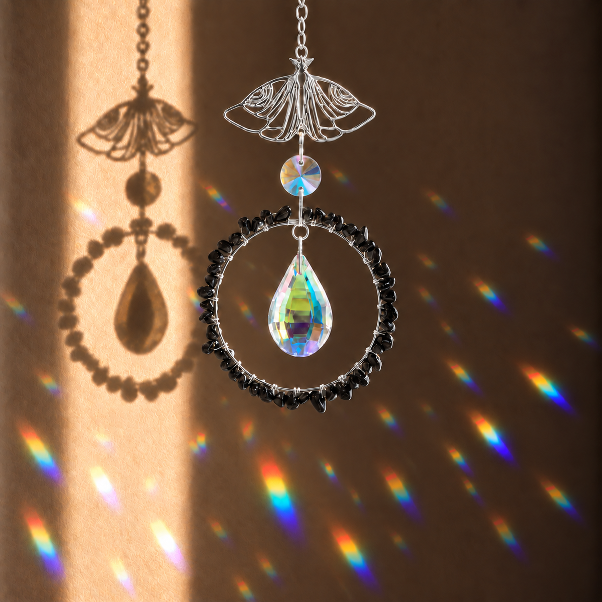 A decorative hanging ornament with a silver butterfly wire frame, a clear teardrop crystal, a black beaded circular ring, and a small iridescent bead, shown against a warm brown wall with rainbow light projections.