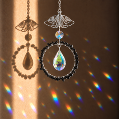 A decorative hanging ornament with a silver butterfly wire frame, a clear teardrop crystal, a black beaded circular ring, and a small iridescent bead, shown against a warm brown wall with rainbow light projections.
