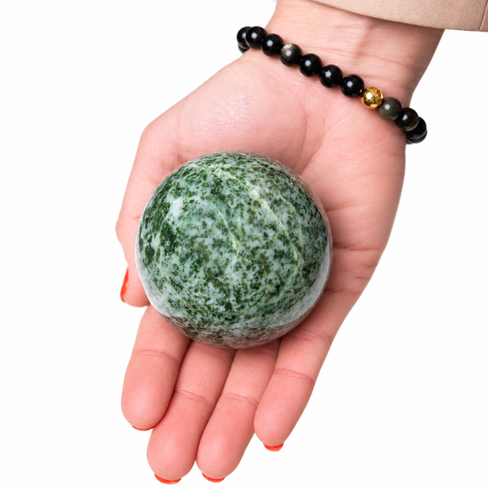 A hand holding a polished green and white marbled stone sphere, with a black beaded bracelet worn on the wrist, against a plain white background.