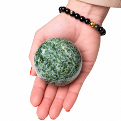 A hand holding a polished green and white marbled stone sphere, with a black beaded bracelet worn on the wrist, against a plain white background.