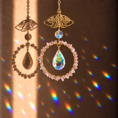 A gold suncatcher ornament with a teardrop-shaped iridescent crystal and a ring of small rose quartz crystals, shown hanging against a brown wall with sunlight casting rainbow reflections.