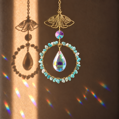 A gold-toned hanging ornament with a butterfly-shaped top, a circular ring of light blue faceted beads, and a large iridescent teardrop crystal in the center, shown against a warm brown background with sunlight and rainbow reflections.