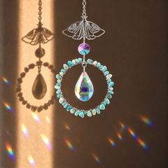 A decorative suncatcher with a silver butterfly-shaped top, a ring of small light blue beads, a large iridescent teardrop crystal, and a smaller matching crystal, shown hanging against a brown wall with sunlight casting rainbow reflections and a butterfly shadow.