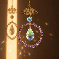 A decorative hanging ornament with a gold-tone top piece, a circular ring set with purple crystals, and a large iridescent teardrop crystal in the center, shown against a warm brown background with sunlight and rainbow reflections.