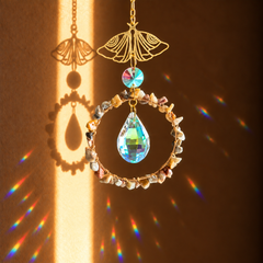 A gold-toned butterfly-shaped suncatcher with a circular ring of small stones and a faceted iridescent teardrop crystal, shown hanging in warm sunlight that casts rainbow reflections and a butterfly shadow on a brown wall.